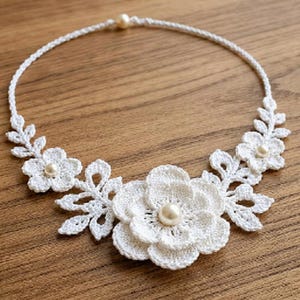 May include: A white crochet necklace featuring a central large floral motif, two smaller side flowers, and delicate leaf patterns. The design is accented with three cream-coloured pearls, one at the centre of each flower, and a single pearl at the clasp.