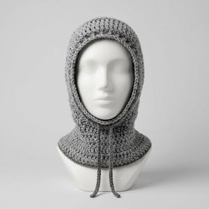 Pode incluir: Uma balaclava de crochê cinza-claro com um padrão de malha texturizada, formato de cabeça arredondado e dois cordões finos de crochê combinando que pendem da área frontal do pescoço. A peça é exibida em um manequim branco liso.