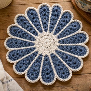 Peut inclure: Un napperon au crochet en forme de fleur, composé d'un cercle central de couleur crème entouré de douze pétales allongés bleu foncé. Chaque pétale est bordé d'une bordure couleur crème, créant un contraste net avec le fil bleu.