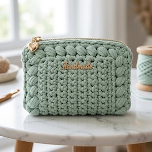 May include: A sage green crochet pouch featuring a thick braided border, a gold-toned zipper with a heart-shaped charm, and a gold metal tag with the word Handmade centered on the front. The pouch is constructed from thick cord yarn.