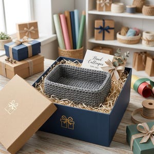 May include: A rectangular dark gray crocheted basket sits inside a large navy blue gift box filled with shredded paper filler. A white card with the text Hand Callrapes FOR YOU and a small sprig of eucalyptus are tucked into the box.