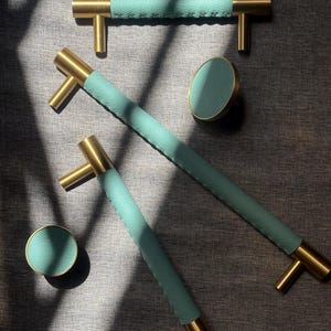 Peut inclure: Un ensemble de quincaillerie pour meubles comprenant deux longues poignées en T et deux boutons ronds. Chaque pièce présente une finition en métal doré brossé associée à un revêtement texturé vert menthe avec des coutures croisées visibles.