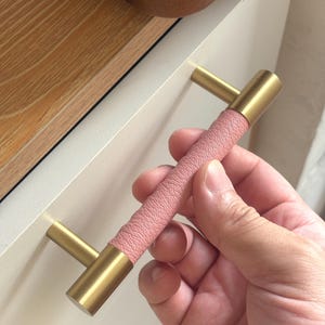 May include: A cabinet handle featuring a cylindrical brass-toned metal bar with a textured, light pink leather-like wrap around the center section, attached to two cylindrical brass-toned metal posts for mounting onto a flat surface.