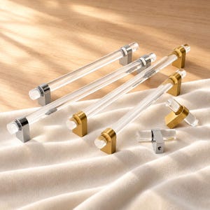 May include: A collection of cabinet hardware including several long, clear acrylic bar pulls and two smaller knobs. The hardware features metallic mounting bases in both polished silver-tone and brushed gold-tone finishes, with clear cylindrical bars.