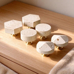 Può includere: Un set di sei pomelli per mobili con piani in pietra travertino beige naturale dalle texture porose. Ogni pomello è montato su una base in ottone lucido, con design esagonali, circolari e a forma di piramide.