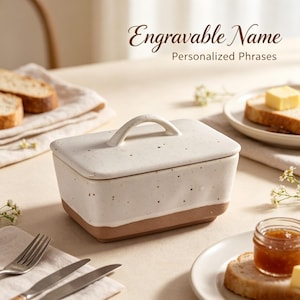 May include: A rectangular ceramic butter dish with a speckled white glaze on the top half and a natural terracotta base. The lid features a curved handle. The text "Engravable Name" and "Personalized Phrases" is visible above the butter dish.