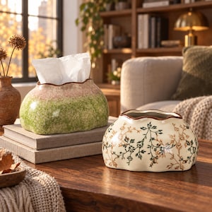 May include: Two ceramic tissue box covers with scalloped edges. One cover features a mottled green and pink finish with white tissues protruding. The second cover is cream-coloured with a delicate brown and green botanical vine pattern printed on it.