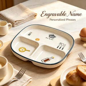 May include: A white ceramic divided plate with rounded corners and a dark blue rim. The plate features colorful illustrations of a frying pan with an egg, a cooking pot, a bottle, and cutlery. Text on the plate reads "Bon Apétit".