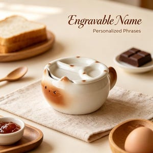 Könnte beinhalten: Eine weiße Keramikschale mit katzenförmigem Design, braunen Akzenten an Ohren und Gesicht sowie einem passenden Deckel mit dreiteiliger Unterteilung. Der Text Engravable Name Personalized Phrases ist über der Schale zu sehen.