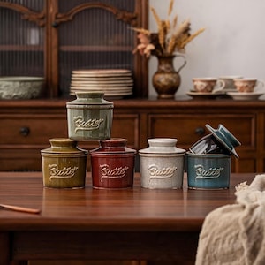 May include: A collection of five ceramic butter crocks in various colors including olive green, mustard yellow, deep red, speckled cream, and teal. Each cylindrical container features a matching lid and the word Butter embossed in a script font.