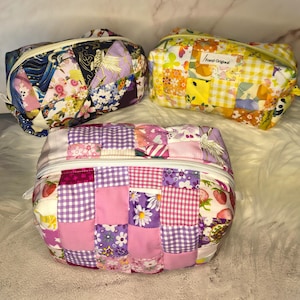 Könnte beinhalten: Drei rechteckige Reißverschlusstaschen aus gestepptem Patchwork-Stoff. Eine Tasche zeigt dunkelblaue und violette Blumenmuster, eine weitere gelb-weißes Vichy-Karo mit Blumenmotiven und die dritte präsentiert florale Muster in Rosa und Violett.