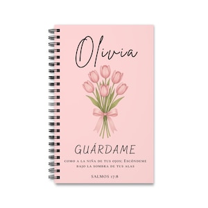 Op de afbeelding: Een spiraalgebonden notitieboek met een lichtroze kaft, voorzien van de naam Olivia in zwarte schrijfletters, een boeket roze tulpen met een strik en de Spaanse tekst GUÁRDAME, COMO A LA NIÑA DE TUS OJOS; ESCÓNDEME BAJO LA SOMBRA DE TUS ALAS, SALMOS 17:8.