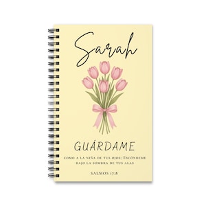 Op de afbeelding: Een spiraalgebonden notitieboek met een lichtgele kaft met een boeket roze tulpen vastgebonden met een roze lint. De naam Sarah staat bovenaan. Onder de bloemen staat de tekst GUÁRDAME COMO A LA NIÑA DE TUS OJOS; ESCÓNDEME BAJO LA SOMBRA DE TUS ALAS SALMOS 17:8.