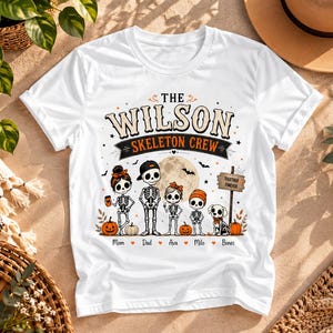 May include: A white t-shirt featuring a graphic of five skeletons and a dog with the text THE WILSON SKELETON CREW. The design includes a full moon, bats, pumpkins, and a sign reading TOGETHER FOREVER, with the names Mom, Dad, Ava, Milo, and Bones below.