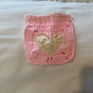 May include: A square crochet patch featuring a light pink border and a central cream-colored heart motif. The piece is constructed with visible yarn stitches, creating a textured, handmade square design with a soft color palette throughout.