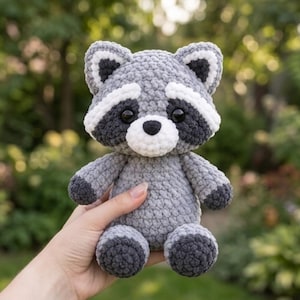 May include: A small, handmade crochet raccoon plush toy featuring a soft, textured grey body, a white muzzle, and dark grey patches around its black eyes. The raccoon has pointed ears with white inner details and dark grey paws and feet.