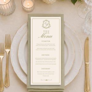 May include: A rectangular paper menu with gold text and a monogram at the top, resting on a sage green folded napkin atop a white plate. The menu lists starters, entrees, and desserts including whipped burrata, chicken breast, and lemon tarts.