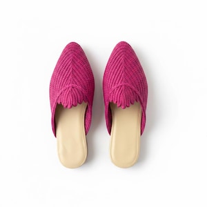 May include: A pair of bright magenta mules featuring a woven, textured upper with a pointed toe and a scalloped edge detail across the vamp. The interior footbed is a smooth, light beige color, providing a contrast to the vibrant exterior.