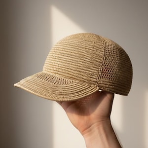 Peut inclure: Une casquette de baseball beige fabriquée dans une matière tissée semblable à de la paille. La casquette présente une calotte arrondie et une visière rigide et incurvée. La texture est uniforme et dense, avec un tissage détaillé et complexe.