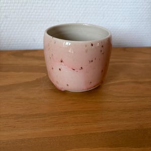 Peut inclure: Une petite tasse en céramique arrondie avec un vernis extérieur rose pâle présentant de subtiles taches sombres. L'intérieur de la tasse est recouvert d'un vernis lisse de couleur crème claire, et le bord est circulaire avec une forme légèrement irrégulière.