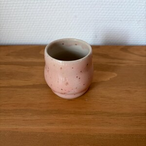 Peut inclure: Une petite tasse en céramique arrondie avec un vernis rose clair et des taches brun foncé subtiles sur tout l'extérieur. La tasse présente une silhouette lisse et incurvée avec une section centrale légèrement plus large et un bord intérieur clair.