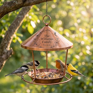 May include: A copper-colored metal bird feeder with a conical roof featuring an engraved floral wreath and the text The Williams Family Est. 2026. The feeder has a circular tray filled with birdseed and is occupied by four small birds.