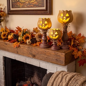 May include: Three jack-o'-lantern candle holders in varying heights, each featuring a glowing yellow face with cut-out eyes and a mouth. The holders have dark brown, turned wooden pedestals tied with twine, arranged among autumn-coloured leaves.