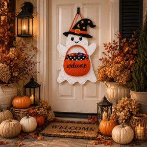 Puede incluir: Un colgante de pared de fantasma acolchado blanco con un sombrero de bruja negro con estrellas amarillas y una luna creciente, que sostiene una cesta naranja en forma de calabaza con la palabra welcome en negro y tela de lunares.