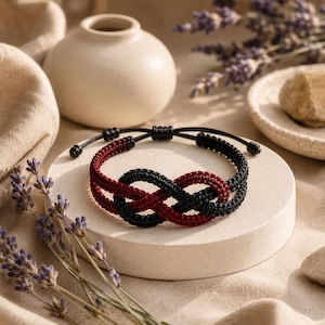 May include: A woven cord bracelet featuring a central infinity knot design. The bracelet is constructed from alternating black and deep red cord segments, with adjustable sliding knots at the back to secure the fit around the wrist.