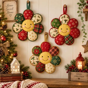 May include: Three quilted flower-shaped potholders with smiling yellow faces, featuring red, green, and cream fabric petals with holiday patterns like snowflakes, holly, and plaid. Each potholder has a fabric loop for hanging on a wall.