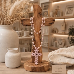 May include: A light pink crocheted rosary featuring a detailed cross pendant, draped over a natural wood cross stand. A small rectangular card sits nearby with the printed text: HANDMADE WITH PRAYER, MADE TO INSPIRE, and a small cross symbol.