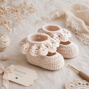 May include: A pair of cream-coloured crocheted baby booties featuring a decorative scalloped ruffle around the ankle opening. The booties are crafted with a dense stitch pattern and include a tag with the text Handmade with love and a heart.