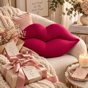 Può includere: Un cuscino a forma di labbra color magenta brillante con una superficie morbida e testurizzata. Il cuscino è posizionato su un divano color crema accanto a un regalo avvolto in carta marrone con un nastro di raso rosa e un'etichetta Handmade with Love.
