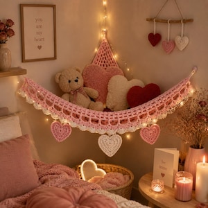 Op de afbeelding: Een roze gehaakte hoekhangmat met een geschulpte rand en hangende hartvormige accenten. De hangmat bevat een beige teddybeer en drie pluche hartvormige kussens in roze, crème en rood, verlicht door een lichtsnoer.