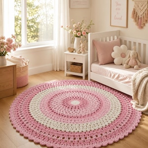 Puede incluir: Una alfombra redonda de ganchillo con anillos concéntricos de hilo rosa suave, crema y blanco. El diseño presenta un patrón de puntos detallado y texturizado con un borde festoneado, creando un motivo circular que cubre el suelo.