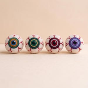 Könnte beinhalten: Vier kugelförmige, nadelgefilzte Wollaugen in einer horizontalen Reihe. Jeder weiße Ball zeigt rote gestickte Adern und eine iris in einer anderen Farbe: Blau, Grün, Rot und Violett, jeweils mit einer schwarzen Pupille in der Mitte.