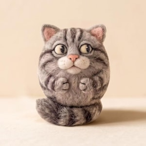 May include: A round, needle-felted cat figurine with grey and white tabby stripes. The cat has large, wide-set eyes, a small pink nose, and white muzzle fur. Its front paws are tucked near its chest, and a thick, striped tail curls at the base.