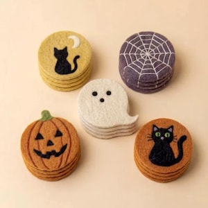 Op de afbeelding: Een set van vijf viltwollen onderzetters met Halloween-ontwerpen: een gele onderzetter met een zwarte kat en maan, een paarse met een wit spinnenweb, een witte in de vorm van een spook, een oranje pompoen en een oranje kattenonderzetter.