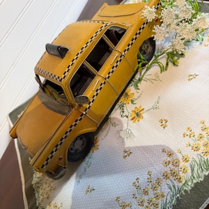 Peut inclure: Un taxi jouet en métal jaune avec une bande à damier noir et blanc sur les côtés, un panneau de toit noir portant l'inscription 5331, des roues gris foncé et un intérieur détaillé visible à travers les fenêtres ouvertes du véhicule.