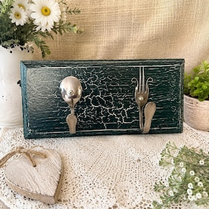 May include: A rectangular wall-mounted hook rack featuring a dark green crackle-finish wooden base. Two silver-toned metal hooks are attached to the front, each fashioned from repurposed cutlery, specifically a spoon and a fork, secured with screws.