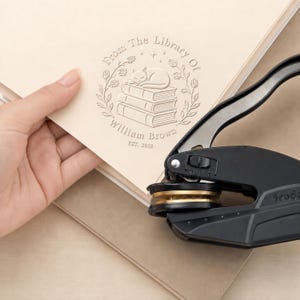May include: A black metal handheld embosser tool creating a circular impression on cream-colored paper. The embossed design features a sleeping cat atop a stack of three books, surrounded by a floral wreath with the text From The Library Of William Brown EST. 2018.