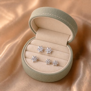 Puede incluir: Dos pares de pendientes de botón en forma de estrella de plata con piedras facetadas transparentes en un joyero redondo cubierto de tela verde salvia con forro interior color crema y una tapa abatible abierta que muestra los cuatro pendientes.