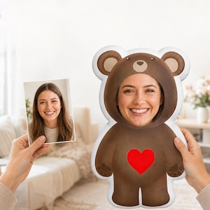 Puede incluir: Un cojín de peluche personalizado con forma de oso marrón y un gran corazón rojo en el pecho. El cojín presenta un recorte circular en el centro de la cabeza del oso, lo que permite que el rostro de una persona sea visible a través de él.