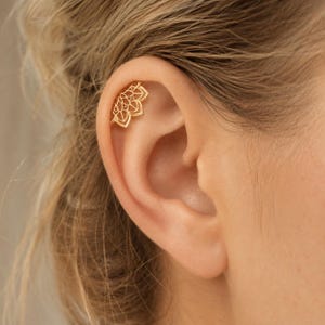 Puede incluir: Una pieza de joyería para piercing de cartílago de oreja en tono dorado con un diseño de abanico semicircular y patrones intrincados de pétalos recortados. La joya está colocada a lo largo del borde superior del cartílago, con un acabado metálico pulido.