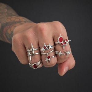 Puede incluir: Una colección de anillos plateados usados en los dedos, con motivos en forma de estrella, diseños de cruces puntiagudas y piedras rojas incrustadas. Los anillos exhiben varias texturas metálicas, incluyendo superficies lisas y detalles.