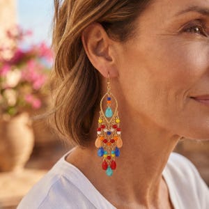 Puede incluir: Un pendiente tipo candelabro en tono dorado con un marco en forma de lágrima adornado con cuentas multicolores en tonos azul, rojo, naranja, amarillo y blanco. El diseño incluye cuentas redondas y en forma de lágrima colgando de finas cadenas.
