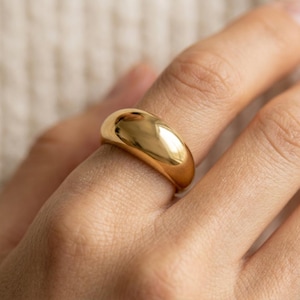 Puede incluir: Un anillo tipo domo en tono dorado pulido con una superficie lisa y redondeada y una banda gruesa y sólida. El anillo presenta un acabado metálico de alto brillo que refleja la luz en su exterior curvo, creando una forma circular.
