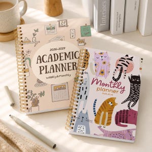 Puede incluir: Dos agendas de espiral con encuadernación de alambre dorado. La agenda académica más grande es de color rosa claro con ilustraciones de líneas y el texto 2026-2027 ACADEMIC PLANNER. La agenda mensual más pequeña muestra ilustraciones de gatos coloridos y el texto Monthly planner 2026-2028.