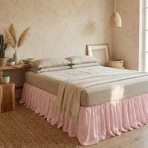 May include: A bed featuring a light pink ruffled bed skirt, topped with beige linen bedding, two matching pillows, and a folded beige throw blanket. The bed skirt has a gathered, pleated design that extends to the floor, covering the base.
