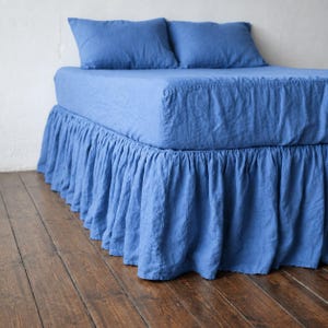 May include: A blue bed set featuring a fitted sheet and a ruffled bed skirt that extends to the floor. Two matching blue rectangular pillows are placed at the head of the bed. The fabric has a visible woven texture throughout the entire bedding set.
